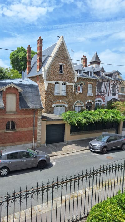 This is a vertical, eye-level, medium shot of a street in Fontainebleau, France, with a focus on two parked cars and several houses. The houses are varied in architectural style, with one featuring a stone facade and prominent chimneys, another with a red brick exterior and a dormer window, and further down the street, houses with half-timbered elements. A black metal fence runs along the bottom of the frame, partially obscuring the foreground. Two grey cars are parked on the asphalt street, one on the left and another on the right. A dark garage door is visible in the center of the image. Lush green foliage cascades over a wall behind the garage. The sky is blue with scattered white clouds.
