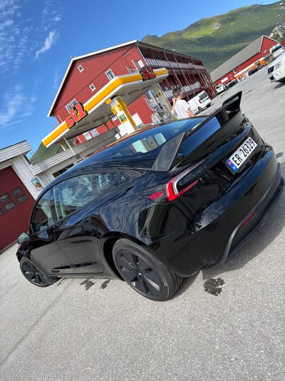 A black Tesla Model 3 is parked on asphalt in front of a red building with a yellow and white awning. The building appears to be a gas station, with the "OK" logo visible on the awning. Behind the Tesla, a white van and other vehicles are parked. Further in the background, a large mountain covered in green vegetation rises against a clear blue sky with a few scattered clouds. The sun is shining, suggesting a daytime scene. The license plate on the Tesla is visible and reads "ER 26329 TESLA.COM N". A person is visible through the car's window, and another person is standing near the gas pumps. The overall scene depicts a stop at a gas station in a scenic, mountainous area.