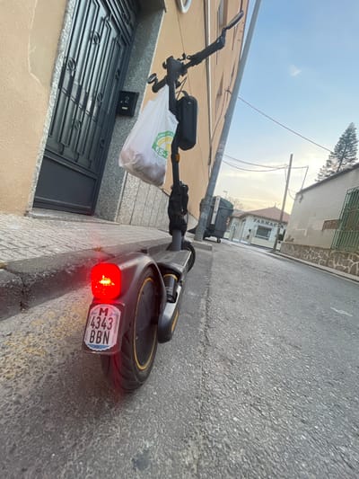 An electric scooter is parked on a street in Cartagena, Spain, with its brake light illuminated. The scooter is positioned facing away from the camera, with its license plate visible. The license plate reads "M 4343 BBN". A white plastic bag hangs from the handlebars of the scooter. The street is paved and appears to be in a residential or commercial area, with buildings on either side. The building on the left has a beige facade and a dark metal door. The building on the right has a white facade with stone accents. In the distance, a building with the word "FARMACIA" is visible. The sky is clear with some clouds.