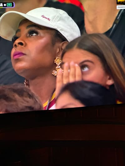 The image shows a close-up of a woman wearing a white baseball cap with "España" embroidered on it in a dark red color. She has on a bright pink lipstick and large, ornate gold earrings shaped like flowers. To her right, another person, possibly a younger woman, is partially visible, covering her face with her hands and looking upwards. The background is dark and blurred, suggesting a crowded or dimly lit environment, possibly a stadium or event. There are no identifiable landmarks that would suggest the image was taken in Bykovo, Russia.