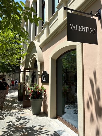 The image depicts the entrance to a Valentino store located in La Roca del Vallès, Spain. A black sign with "VALENTINO" in white lettering hangs above the entrance. To the left of the entrance, a large potted plant with red flowers is situated on the pavement. The building features a light pink facade with arched entrances and windows. Sunlight casts dappled shadows from overhead trees onto the grey tiled pavement. Several people are walking past the store, with one individual in the foreground wearing a leopard print skirt. The environment appears to be an outdoor shopping area.