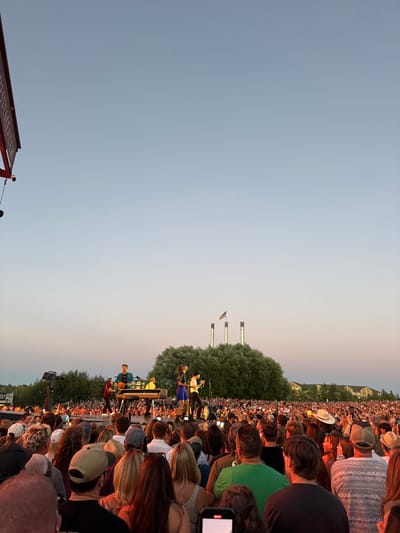 The image captures an outdoor live concert during twilight at the Hayden Homes Amphitheater in the Old Mill District of Bend, Oregon, United States. An elevated stage is positioned centrally, featuring a band of approximately five individuals. One performer is seated at a keyboard, while others stand, some holding stringed instruments. The immediate foreground is densely populated by a large crowd of attendees, primarily viewed from behind, facing the stage. Several individuals within the crowd are holding smartphones aloft, seemingly recording the performance. In the background, behind the stage, a large, rounded cluster of trees is prominent, with three industrial chimneys and an American flag visible above the tree line. Distant buildings are discernible on the right horizon. A partial red sign, likely part of the venue's branding, is visible in the upper left corner. The sky displays a gradient from light blue at the zenith to a faint pinkish-orange hue near the horizon, consistent with sunset.