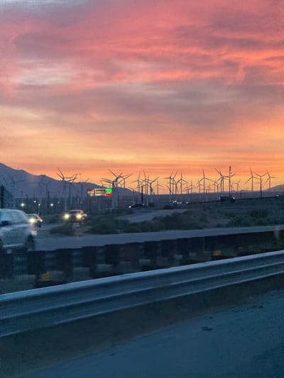 This image captures a scenic sunset over a desert landscape, likely near Desert Hot Springs, United States. The sky is illuminated with vivid hues of pink, orange, and light purple, indicative of twilight. Scattered clouds stretch across the expansive sky, painted by the fading sunlight.
In the mid-to-far ground, numerous wind turbines are silhouetted against the colorful horizon, their distinct shapes suggesting a vast wind farm. Mountains or hills are faintly visible in the distance beneath the sky. A highway occupies the middle ground, where several cars are visible. Some vehicles have their headlights on, appearing as bright blurs against the diminishing light, suggesting movement. One car is partially visible in the left foreground, also in motion.
A green and white roadside sign is discernible in the midground, featuring the word "Pilot" in white letters on a green background, likely indicating a "Pilot" travel center or gas station. The immediate foreground is dominated by a metallic highway guardrail, implying the photograph was taken from a vehicle or from the roadside. The scene conveys a sense of travel through an energy-producing desert environment during a beautiful sunset.