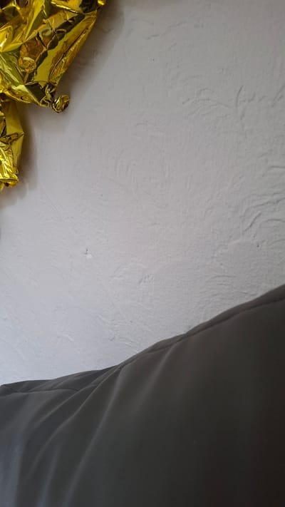 The image shows a close-up, angled view of a gold foil balloon and a dark grey fabric surface against a textured white wall. The balloon is crinkled and appears to be a solid piece. The fabric surface is smooth and forms a curved shape, suggesting it might be part of a piece of furniture or clothing. The white wall has a rough, bumpy texture. There are no visible people or any indication of the city of Dachi, Kazakhstan.
