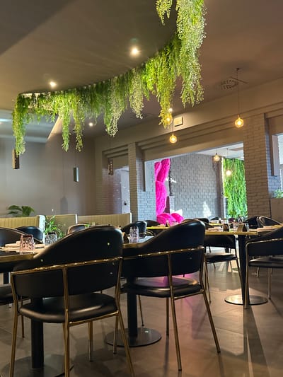 The image depicts the interior of a modern restaurant, likely in Ponteterra, Italy, featuring an abundance of hanging greenery. The ceiling is adorned with lush, artificial green vines that cascade downwards, creating a vibrant, organic aesthetic. This is complemented by a vertical green wall in the background, further enhancing the biophilic design.
The restaurant is furnished with black leather chairs that have sleek, gold-colored metal frames. These chairs surround dark, reflective tables, set with plates, glasses, and cutlery. A few small bottles, possibly soy sauce or condiments, are visible on one of the tables. The lighting appears to be a mix of recessed ceiling lights and decorative pendant lamps with warm bulbs, casting soft glows.
The environment suggests a contemporary dining space, possibly catering to Asian cuisine, given the presence of what looks like soy sauce bottles. The background reveals glimpses of a pink, fluffy decoration and exposed brickwork, adding texture and visual interest to the space. No people are visible in the image, and the scene is static, focusing on the restaurant's decor and arrangement. The floor is tiled in a neutral tone.