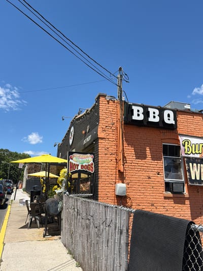 The image depicts the exterior of a restaurant with orange brick walls and a black facade on the upper section. A large sign reading "BBQ" in white block letters is mounted on the black facade. Another sign with a hot dog graphic reads "Nathan's HOT DOGS". To the right of the "BBQ" sign, a sign with "Burgers" in yellow letters and "W" in white letters are visible.

The restaurant has an outdoor seating area with yellow umbrellas and tables with chairs. A large charcoal grill is positioned near the seating area. A tall fence made of vertical wooden slats runs along the sidewalk in front of the restaurant, with string lights attached to it. A black mesh mat is draped over a chain-link fence to the right.

The scene appears to be on a sidewalk next to a street, with parked cars visible on the left. The sky is blue with some scattered white clouds. The image is taken from a low angle, looking up towards the restaurant and the sky. The location is specified as Edgewater, United States of America.