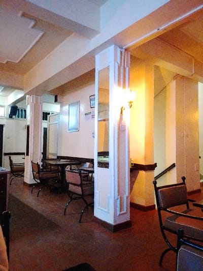 The image depicts an interior scene, likely a common area such as a cafe, restaurant, or hotel lobby. The space appears unoccupied or very quiet.
The setting is characterized by warm, inviting tones. The walls and ceiling are predominantly light-colored, ranging from cream to off-white. The ceiling features multi-leveled sections and decorative geometric beams, suggesting an older architectural style, possibly with Art Deco influences.
Several round tables with dark, possibly black, metal bases are arranged across the floor. Accompanying these are chairs with black metal frames and light-colored upholstered seats and backs. One chair is partially visible in the foreground on the right.
Prominent architectural features include several columns or pillars. A large, central pillar stands out with its light-colored panels and a reflective surface, likely a mirror, on one side, which reflects parts of the room, including tables and chairs. Attached to this central pillar is a wall sconce emitting a bright, warm, yellowish light, which casts a glow on the adjacent wall and pillar. Another similar pillar is visible further back on the left. The bases of these pillars have a darker trim.
In the background, multiple doorways are visible, suggesting access to other rooms or sections of the building. On the far wall, there are two framed items, possibly pictures or notices. Between these frames, a small, rectangular sign with some text is visible, but the text itself is too small and blurry to be deciphered.
To the far right, a dark banister and part of a staircase are visible, indicating a change in floor level. The floor throughout the area is a dark reddish-brown, possibly polished concrete or large tiles, showing some texture and wear.
The overall lighting in the room is a mix of the warm light from the sconce and ambient light, with some cooler, possibly bluish, tones visible in the upper left section of the ceiling, perhaps from an unseen window or different light fixture. No people are present, lending a serene and quiet atmosphere to the scene.
