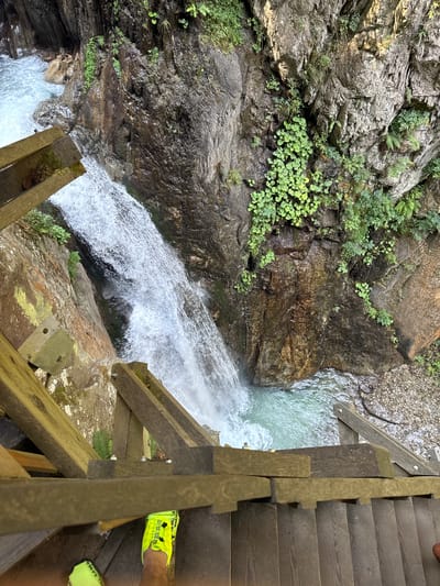 This is a first-person perspective, downward-facing photograph of a wooden staircase leading towards a cascading waterfall within a rocky gorge. The viewer's neon yellow athletic shoes are visible at the bottom of the frame, suggesting the photographer is standing on the stairs. The waterfall plunges into a pool of turquoise water at the bottom of the gorge. The rock faces of the gorge are covered in patches of green vegetation, including ferns and circular-leafed plants. The staircase is constructed of weathered wooden planks and railings, some of which appear to be reinforced with metal brackets. The scene is outdoors, likely in a natural park or hiking area. This image was taken in the city of Martigny-Combe, Switzerland, at the Gorge de la Tine.