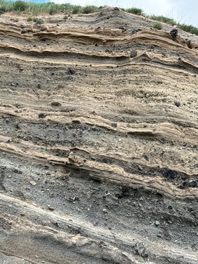 The image displays a close-up view of a rock formation, characterized by distinct horizontal layering. The strata vary in color, with bands of light tan, gray, and darker shades of gray and black. These layers are composed of smaller rock fragments, pebbles, and volcanic ash, suggesting a history of deposition and erosion. Some larger, dark-colored rocks are embedded within the layers. Sparse green vegetation, appearing as grass and small bushes, grows along the top of the formation. The sky is visible in the upper background, appearing overcast with a pale blue hue peeking through. The scene is static, with no visible people or animals. The environment appears to be an outdoor geological outcrop. Based on the provided information, this rock formation is located in the city of Tsghuk, Armenia.