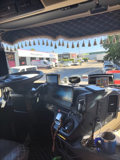 The image shows the interior of a truck cabin, looking out through the windshield. The dashboard features a steering wheel, multiple screens, and various controls. A beaded necklace hangs from the dashboard, and a phone rests on top of it. Two religious icons are also visible. Outside the truck, a street scene in Saint-Herblain, France, is depicted, with buildings, parked cars, and trees under a blue sky. The building directly in front of the truck has signage that reads "Pneus & Entretien" and "PROFIL."