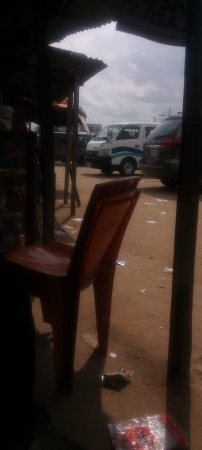 The scene is taken from inside a small, shaded structure, likely a shop or stall, in Elelenwa, Nigeria, located at latitude 4.856 and longitude 7.068. The structure has a corrugated metal roof and wooden support beams. A single, brown plastic chair is positioned in the center of the scene, partially obscured by the wooden support beam. The chair faces away from the viewer. Two vehicles are visible in the background. The vehicle on the left is a white minivan with a blue and green stripe and the number "002" displayed on its side. The vehicle on the right is a dark, possibly black, SUV with a partially obscured license plate. Both vehicles appear to be parked. The ground is a dusty, light brown color with various pieces of trash scattered across its surface. The view is partially obscured by the support beam, suggesting that the viewer is taking the picture from within the shaded structure. The photo captures the scene from a low angle, slightly angled upward toward the vehicles. There are no people present in the scene.