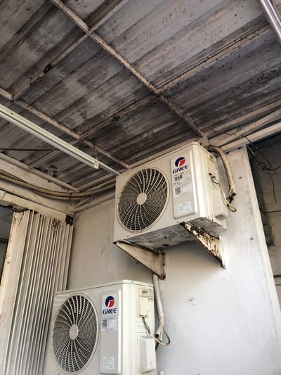 Two white Gree brand air conditioning outdoor units are mounted on a white textured wall beneath a weathered metal roof structure. The upper unit is mounted on a rusty, L-shaped metal bracket. The lower unit is mounted directly onto the wall. Both units have visible circular grilles and labels with the Gree logo. Electrical conduits and wires are routed along the wall and up towards the ceiling. A section of white corrugated metal siding is visible on the left side of the image. The ceiling appears to be constructed of corrugated metal panels supported by white metal beams, with some signs of rust and wear. The overall scene suggests an outdoor or semi-enclosed space, possibly a balcony or patio, located in Putat, Indonesia.