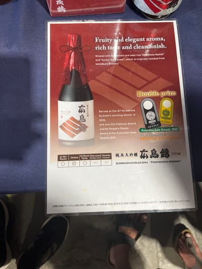 The image is of a restaurant menu, likely a Japanese restaurant. The menu features a bottle of Junmai Daiginjo-shu "Hiroshima-Nishiki" sake. The bottle is surrounded by information about the sake, including its aroma, taste, and finish. It is described as having a fruity and elegant aroma, rich taste, and a clean finish. The sake was brewed with the "Hiroshima Nishiki" rice and "kyokai No. 7 yeast." The menu mentions that this sake was served at the G7 Hiroshima Summit's working dinner in 2023 and won the Platinum Award and the People's Choice Award at the Australian Sake Awards 2023. The menu also includes temperature recommendations for drinking the sake. The menu is on a table, and the bottom of two people's feet, wearing sandals, can be seen. The location is in Hamilton, Australia, based on the coordinates (-27.439, 153.075), which corresponds to a residential area with a mix of housing and businesses.