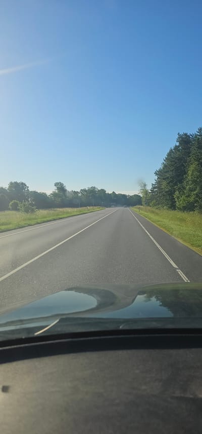 The image shows a view from inside a vehicle driving on a two-lane asphalt road. The road is bordered by green grass and trees on both sides. The sky is clear and blue. The foreground shows the dashboard and hood of the vehicle, with a reflection of the road and sky visible on the windshield. The road stretches into the distance, with a white line marking the lanes. There are no other vehicles immediately visible in front or behind. The scene appears to be in a rural or suburban area. The location is specified as Kersalu, Estonia.