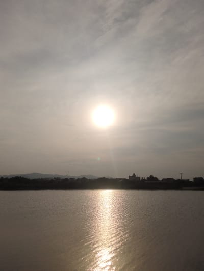 The image depicts a serene sunset over a body of water, likely a lake or river, in Dao Xa, Vietnam. The sun, a bright, diffused orb, hangs in the hazy sky, casting a shimmering reflection on the water's surface. The water is calm, with gentle ripples catching the light, creating streaks of gold. In the distance, a silhouetted treeline and low-lying hills form the horizon. Above the treeline, a few buildings are visible, including what appears to be a church with a dome. The overall atmosphere is tranquil and peaceful, characteristic of a rural sunset. There are no people or animals visible in the frame, and the focus is on the natural landscape and the interplay of light and water. The sky is overcast, with thin clouds softening the sunlight.