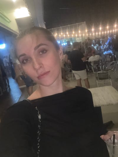 A woman is taking a selfie outdoors at night, with a crowd of people in the background. The scene appears to be a busy outdoor gathering or bar, possibly at the Jersey Shore, given the lights and the general atmosphere. The woman is wearing a black top and has her hair pulled back. The background is filled with people mingling and sitting at tables, illuminated by string lights. The location is identified as Seaside Heights, United States of America.
