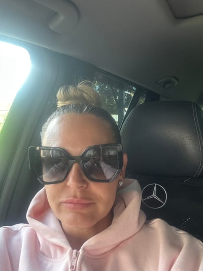 A woman is shown in a close-up shot, seated in the passenger seat of a car. She is wearing large, dark-rimmed sunglasses and a light pink hoodie. Her hair is pulled up in a messy bun. The seat behind her appears to be black leather with white stitching and has a Mercedes-Benz logo embroidered on it. The interior of the car is dark, but sunlight streams in from the right side of the frame, suggesting it is daytime. The woman is looking towards the camera, and her expression is neutral. There are reflections in her sunglasses, showing a glimpse of the outside environment, which appears to be greenery. There is no visible text in the image. The overall impression is a casual selfie taken during the day.