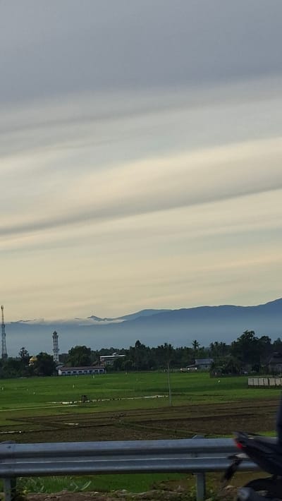 The image captures a landscape view of Mukee Gogo, Indonesia. In the foreground, a silver metal guardrail runs horizontally across the bottom of the frame, with a portion of a dark motorcycle visible on the right. Beyond the guardrail, a field of green crops, possibly rice paddies, stretches into the distance. The field is interspersed with some tilled brown earth. In the middle ground, a line of trees borders the field, behind which are several buildings. A prominent, tall, white minaret of a mosque stands out among the trees on the left, accompanied by other structures including what appears to be a school building. Further in the background, a range of blue-tinted mountains is visible, partially shrouded in a layer of low-lying clouds or mist. The sky above is a muted gray with distinct horizontal cloud formations, suggesting either an overcast day or dawn/dusk lighting. A tall communication tower is also present on the far left of the image, near the mosque.