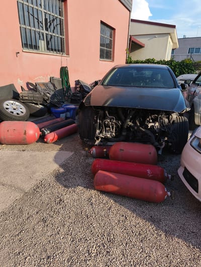 In Chiugiana, Italy, in an outdoor area with a gravel and concrete ground surface, a heavily damaged black car sits in front of a pink building with barred windows. Several large, red cylindrical tanks, some black, are scattered on the ground around the car. In the background, other vehicles and building structures are visible, along with green vegetation and a cloudy blue sky. The scene suggests a junkyard or vehicle repair facility.