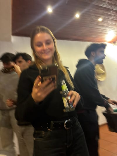 A slightly blurred image shows a woman taking a selfie with her cell phone. She holds a small beer bottle in one hand. A man wearing a black hoodie is behind her and to the right. He seems to be holding a drink as well. Another person is visible below the woman, wearing a gray shirt. The background includes a wooden ceiling with a series of lights and a pale wall.