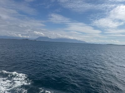 The image depicts a vast expanse of dark blue ocean under a partly cloudy sky. The water is choppy, with white foam and waves visible in the foreground on the left side of the frame, indicating movement. In the distance, a mountainous coastline stretches across the horizon, with some peaks obscured by clouds. The sky is a mix of bright blue patches and white, wispy clouds, with some larger, more defined clouds casting shadows on the distant mountains. The overall impression is one of a rugged, natural landscape. The specific location in Nordsolvær, Norway is a coastal area characterized by its maritime environment, mountainous terrain, and open sea. There are no people or man-made objects visible in the image.