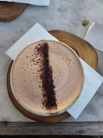 The image displays a top-down view of a light brown, foamy beverage contained within a cup, centrally garnished with a vertical stripe of dark brown powder and scattered dark speckles across its surface. The cup rests on a circular wooden coaster, which partially overlaps a folded white paper napkin. These items are situated on a light-colored, mottled surface, likely a table. In the upper left background, a second, similar circular wooden coaster is visible. To the upper right, a single green pistachio nut lies adjacent to a crumpled piece of white paper. The immediate foreground features a weathered, horizontal wooden surface. No individuals or active interactions are depicted. The scene is located in Ban Sahakon 2, Thailand.