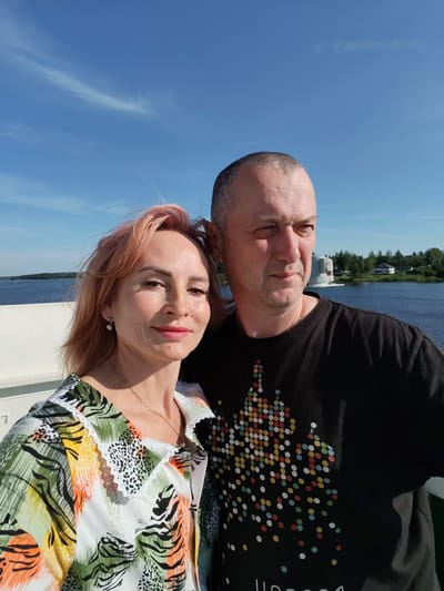 A man and a woman are posing for a selfie on the deck of a boat. The woman, on the left, has reddish-blonde hair and is smiling at the camera. She is wearing a colorful, patterned shirt with a white lace collar and a silver necklace. The man, on the right, has short grey hair and is looking off to the side. He is wearing a black t-shirt with a pattern of colorful dots.
The background shows a body of water, possibly a river or lake, with trees and buildings on the far bank. A large, grey structure is visible in the water to the right of the couple. The sky is bright blue with some wispy white clouds, suggesting it is a sunny day. The lighting indicates it is likely daytime, possibly late morning or afternoon. The overall mood appears relaxed and pleasant. The location is identified as Nova, Russia. No visible text is present in the image.