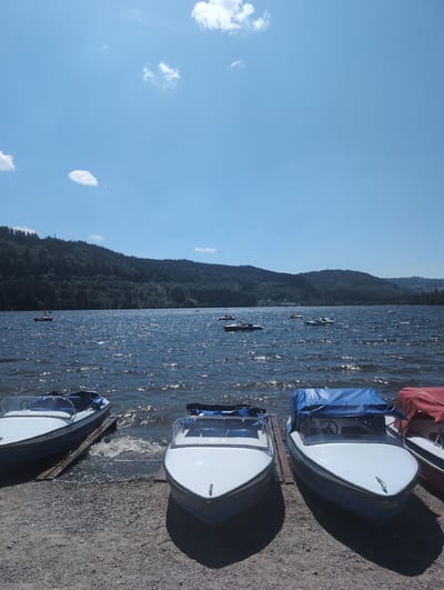 The image depicts a sunny day at Lake Titisee in Titisee-Neustadt, Germany. In the foreground, three motorboats are parked on a gravelly shore. Two of the boats are white with blue interiors, and one is covered with a blue tarp, while another is partially covered with a red tarp. The middle boat is partially submerged in the water, with waves lapping at its hull. The lake is choppy, reflecting the sunlight and creating a shimmering effect. In the background, a dense, tree-covered hillside rises from the opposite shore. Several other boats, including pedal boats, are scattered across the lake, and a few distant buildings are visible along the shoreline. The sky is a clear blue with a few scattered white clouds.