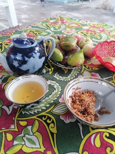 This is a medium shot of a table setting with food and a tea set, likely in an outdoor or semi-outdoor setting in Gulshan, Uzbekistan. The table is covered with a vibrantly patterned cloth featuring red, green, and yellow designs, possibly depicting floral motifs.
In the foreground, there are two bowls. The left bowl contains a light yellow liquid, presumably tea, with a few dark specks floating in it. It has a white interior with a decorative gold rim. To the right, another bowl holds a portion of cooked rice, appearing to be pilaf or a similar dish, with small pieces of vegetables or meat mixed in. A silver spoon rests in this bowl, with some rice on its surface.
Behind the bowls, to the left, is a decorative blue and white ceramic teapot with a matching lid. The teapot has a handle and a spout. To the right of the teapot, on a gold-rimmed plate, is a selection of fruits. These include several green figs, some with reddish blemishes, and two apples, one predominantly red and the other with red and green coloring. A small, red woven basket sits to the far right, with a piece of bread visible inside.
In the background, blurred figures of at least two people are visible standing. The ground appears to be concrete or pavement. The overall scene suggests a meal or a break, possibly in a garden or courtyard area.