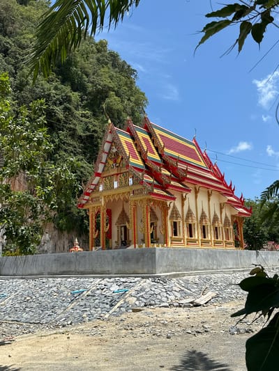 A Buddhist temple is situated in a verdant, hilly environment in Thung Yai, Thailand. The temple features a multi-tiered, ornate roof adorned with vibrant red, yellow, and green tiles, and decorated with intricate golden trim and mythical figures. The facade of the temple is cream-colored with gold accents, including numerous small, arched windows with golden frames. A stone retaining wall and a slope covered in gray stones, with visible blue pipes, lead up to the temple's base. Lush green trees and a rocky cliff face surround the temple, with palm fronds and other foliage visible in the foreground and framing the scene. The sky above is a clear blue with scattered white clouds. There are no people visible in the image.