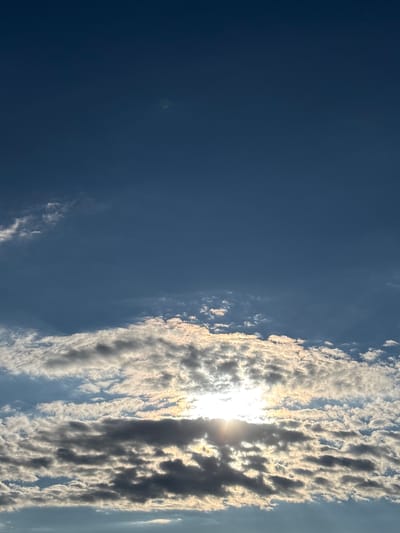 This is a vertical, full-frame, eye-level, outdoor, full-color photograph of the sky above Novaya Usman, Russia. The image captures a dynamic sky filled with a mixture of clouds and sunlight. The upper portion of the image is dominated by a clear, deep blue sky with a subtle lens flare visible as a faint greenish circular artifact near the top center.
The lower half of the image features a dense band of clouds. These clouds exhibit a variety of textures and lighting conditions. Some are bright white and illuminated by the sun, appearing fluffy and ethereal, while others are darker, tinged with gray and casting shadows, suggesting their density and thickness. The sun itself is partially obscured by these clouds, peeking through the breaks and casting rays of light that create a radiant glow and highlight the edges of the cloud formations. The light suggests it is either early morning or late afternoon.
There are no people, animals, buildings, or any other terrestrial objects visible in the frame. The focus is entirely on the atmospheric conditions. The composition draws the viewer's eye from the bright, sunlit clouds upwards into the vast expanse of the blue sky.