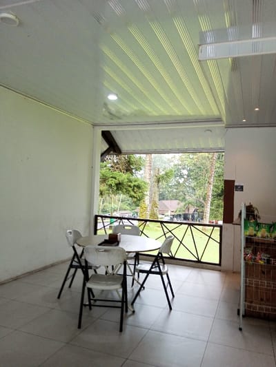 The scene depicts an unoccupied, partially enclosed, open-air dining or seating area located in Pachmarhi, India. The space features a light-colored tiled floor, off-white walls, and a white PVC strip ceiling with parallel grooves and embedded LED lighting. Centrally positioned is a round, light-colored table surrounded by four white plastic folding chairs with black frames. On the table rests a dark wooden tissue box holder and a rectangular holder, possibly for a menu. To the far right, a tall, narrow wooden display rack with wire shelves holds numerous packaged snack items. The open side of the area is bordered by a dark metal railing with a decorative criss-cross pattern. Beyond the railing, a lush green landscape unfolds, dominated by tall trees and a distant, low-slung building with a dark roof and light-colored walls, suggestive of a resort or lodge within a natural setting. No individuals are present, and no actions or interactions are occurring. The lighting suggests an overcast or diffused daylight condition.