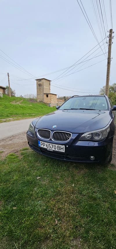 A dark-colored BMW is parked partially on a paved surface and partially on grass. The front of the car, including the grill and license plate reading "PP 6570 BH", is visible. In the background, a concrete and brick structure is present, and overhead power lines are seen. The scene appears to be in an open, rural area, possibly in the vicinity of Pobit Kamak, Bulgaria.