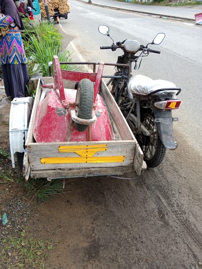 In Geulanggang Baro, Indonesia, a modified motorcycle with a sidecar is parked on the side of a paved road. The sidecar is constructed from wood and contains a red wheelbarrow. A person, partially visible on the left side of the image, is wearing a dark headscarf and a colorful patterned tunic over a dark skirt. Behind the motorcycle, a large pile of yellow produce, possibly corn, is loaded onto another motorcycle. The environment appears to be a rural or semi-urban area, with a dirt shoulder and some vegetation along the roadside. The weather is overcast.