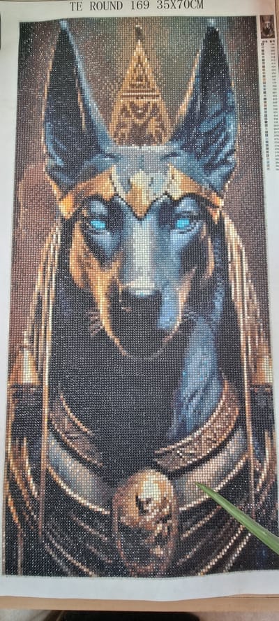 The image displays a diamond painting of an Egyptian god, likely Anubis, characterized by its jackal head and regal adornments. The artwork is created using small, faceted beads that reflect light, giving the image a shimmering quality. The god's eyes are a striking electric blue, contrasting with the darker tones of its fur and the gold and bronze elements of its headdress and ceremonial collar. The background is a textured blend of browns and grays, creating a sense of depth.
The diamond painting is situated on a white surface, and in the lower right corner, a portion of a green leaf or plant stem is visible, extending into the frame. The top edge of the canvas has text printed on it, which reads "TE ROUND 169 35X70CM". To the right of the main artwork, a color chart or legend is partially visible, indicating the various bead colors and their corresponding codes.
There are no discernible elements within the image that would suggest a specific location in the city of Naaldwijk, Netherlands. The context provided is solely that of the diamond painting itself and its immediate surroundings.