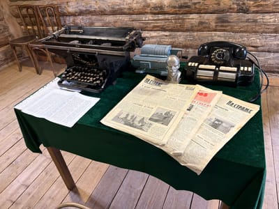 In a room with log walls and a wooden floor, a dark green tablecloth covers a table. On the table are several objects: a black manual typewriter, a light blue mechanical calculator, a silver bust of Lenin, and a black rotary telephone. Scattered across the table are newspapers. Two wooden chairs are visible in the background. The scene appears to be a static display, possibly in a museum or historical exhibit, depicting a historical office setting. The city of Ust'-Kabyrza, Russia is a rural settlement, and this scene may represent a historical office from that region or a similar setting.
