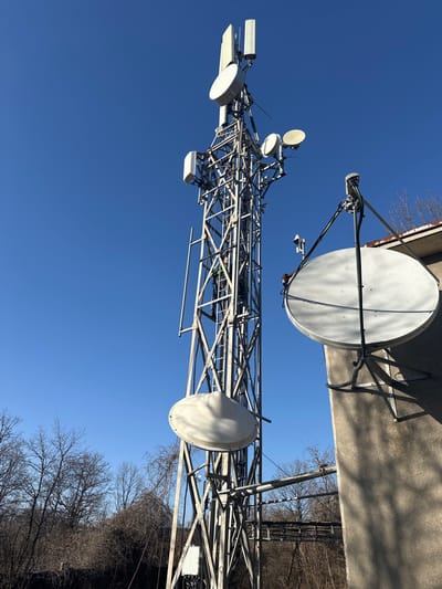 The image depicts a telecommunications site in Opaka, Bulgaria, under a clear blue sky. A galvanized steel lattice tower stands centrally, supporting multiple white rectangular panel antennas and various circular parabolic dish antennas at differing heights. A smaller, white, dome-shaped antenna is also affixed lower on the tower structure. One individual, wearing dark clothing and a green element possibly part of safety gear, is observed actively climbing the tower's internal ladder, positioned approximately one-third of the way up its visible height. To the right, a multi-story building with a textured concrete facade features a large white parabolic satellite dish mounted on its exterior wall via a metal frame. The background consists of bare deciduous trees and shrubs, indicating a dormant season.