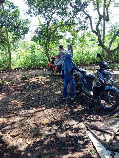 The image depicts an outdoor scene in Bulukandang, Indonesia, under bright daylight conditions, characterized by dappled sunlight and shadows from overhead trees. In the mid-ground, three individuals are present near two motorcycles on an uneven dirt surface.
On the left, a male individual in a light-colored short-sleeved shirt and patterned shorts is seated on a red Honda-branded underbone motorcycle. Behind him, a female individual, dressed in a light-colored hijab and attire, is seated as a passenger. The red motorcycle is facing towards the left of the frame.
To the right, a dark blue or black Honda scooter is parked. A third individual, a female wearing a blue hijab, blue top, and blue jeans, stands partially obscured by the scooter, facing away from the primary group on the red motorcycle. Her back is mostly to the viewer.
The ground consists of exposed soil and small gravel, interspersed with scattered dry leaves and twigs. The background is dominated by dense green foliage, tall trees, and what appear to be banana plants, suggesting a rural or semi-rural environment. A thin wire or cable is visible stretched between trees in the upper portion of the frame. No direct interaction between the standing individual and those on the red motorcycle is evident.