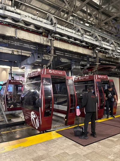 The image shows a series of cable car cabins at the Awana Skyway station in Genting Highlands, Malaysia. The cabins are maroon with white stripes and the logo of "Resort World Genting." One cabin is labeled "87" and another "27" with "Rearth". People can be seen inside some of the cabins. A person is wearing a black jacket with the acronym "STS" on the back cleaning the outside of a cabin. The overhead structure is composed of metal support beams and machinery. The platform is gray and yellow striped.