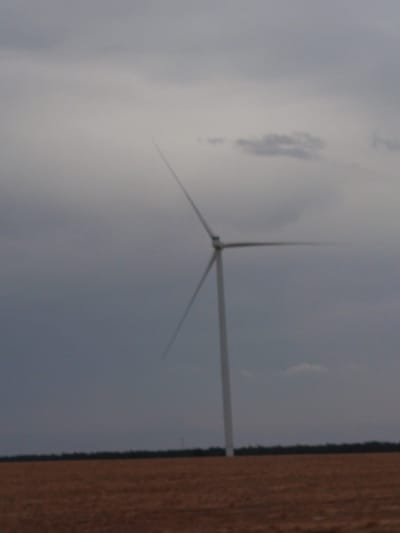 An outdoor, medium-distance shot captures a single wind turbine standing in a flat, harvested field under a gray, cloudy sky. The wind turbine, positioned slightly off-center to the right, features a white tower and three long, angled blades. The field is a uniform brown, suggesting it is dry and has recently been harvested. A dark, blurry line of trees or foliage runs along the horizon, separating the field from the sky. The overall atmosphere is muted and somewhat overcast. There are no people or animals visible in the frame.