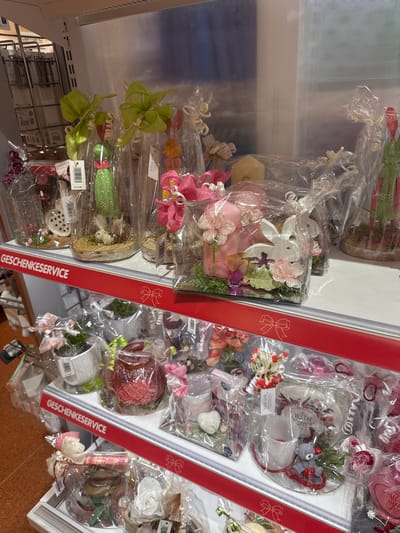 The image shows a selection of wrapped gifts or decorative items on shelves, likely in a store. The items are individually packaged in clear cellophane wrapping with decorative bows and floral accents. A shelf divider is visible, marked with "GESCHENKESERVICE" (German for "Gift Service") in red lettering with a bow motif. Based on the available information, it can be inferred that the location is in Amstetten, Austria.
