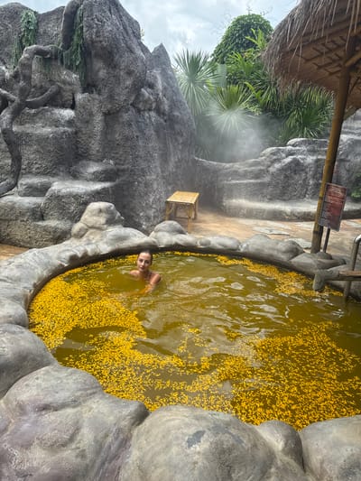 A woman is chest-deep in a round, hot spring pool filled with yellow flower petals, surrounded by artificial rock formations. The pool is located in Ban Khlong Khian, Thailand, based on the location metadata. There is steam rising near a small bamboo chair and thatched shelter. A sign with text is posted nearby.