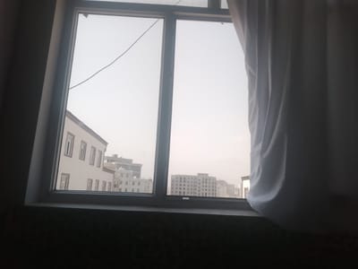 The image shows a view from a window looking out onto a city. The window is divided into two panes by a vertical mullion, and a horizontal mullion divides the top and bottom sections. A white curtain is partially drawn on the right side of the window, obscuring some of the view.
Outside the window, several buildings are visible. On the left, there is a light-colored building with multiple windows. Further in the distance, a row of white buildings with many windows can be seen, and beyond that, larger, more modern buildings with grid-like window patterns. The sky is overcast and light-colored, suggesting it might be early morning or late afternoon, or a cloudy day.
This image is not located in Vahdat, Tajikistan.