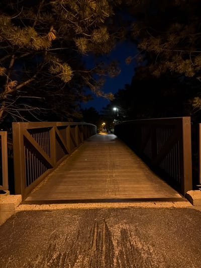 The image depicts a wooden pedestrian bridge illuminated by streetlights at night. The bridge is wide and appears to be made of wooden planks, with dark brown wooden railings on either side. The railings have a lattice design with vertical slats and diagonal braces. The bridge is surrounded by trees, some of which are pine trees with illuminated needles. The sky is dark blue, indicating nighttime. The ground in the foreground appears to be asphalt or a similar dark surface, with some scattered white debris. The location is identified as being in the city of Vernon Hills, United States of America.