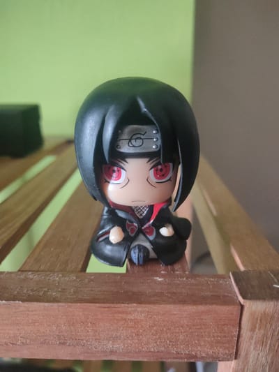 A small, stylized figurine of Itachi Uchiha from the anime series Naruto is positioned on a wooden surface. The figurine is dark-haired, with a black headband featuring a silver forehead protector. Its eyes are red with black pupils, and it wears a dark cloak with red accents and a high collar. The figurine is centrally located in the frame. The background consists of a soft green wall and a blurred brown area. The wooden surface appears to be part of a slatted shelf or bench. No people are visible. The scene is indoors, with soft, natural-looking lighting. The figurine is the sole subject of focus, and no specific location in Figueres, Spain, can be determined from the visual information provided.