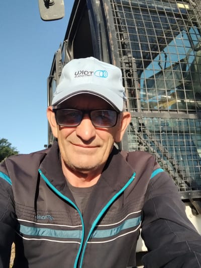 This is a selfie of a man wearing a black jacket with blue and white trim, sunglasses, and a light blue baseball cap. The cap has a logo with the text "UNION" and a circular design. The man has stubble and appears to be smiling. The scene is outdoors with a clear blue sky. The man is positioned in front of a metal grate, which appears to be part of a vehicle's protective cage. In the background, the reflection in the vehicle's window shows a blue sky and possibly trees. The lighting suggests it is daytime, likely morning or afternoon, with bright sunlight creating shadows. The image seems to be taken at a slight upward angle. The man's clothing suggests he might be involved in outdoor work or a recreational activity. The location is indicated as Novoselytsia, Ukraine.