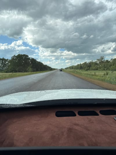 The image depicts a straight asphalt road cutting through a verdant landscape under a heavily clouded sky, viewed from the interior of a vehicle. The scene is situated in Zerendi, Kazakhstan. The sky is dominated by large, grey and white cumulus clouds, with intermittent patches of blue visible. Both sides of the two-lane road are lined with green foliage; dense trees and brush border the left, while a wider expanse of grassy field and scattered smaller trees lies to the right. The road surface appears damp, exhibiting reflections suggesting recent precipitation or moisture. Several vehicles are visible in the far distance, receding along the road. The immediate foreground captures a portion of the vehicle's interior, specifically the dashboard, which is finished in a textured, dark brown material. The lower edge of the windshield shows numerous small water droplets. Three dark, rectangular indentations, possibly vents, are present on the dashboard. No people are discernible in the scene.
