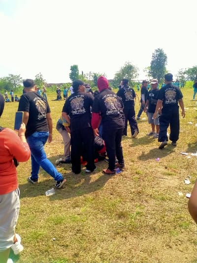 This image depicts an outdoor scene in Celep, Indonesia, on a bright, clear day. The foreground and mid-ground feature a group of approximately ten to fifteen individuals, predominantly adults, gathered on a sparse, dry grassy field with some green patches. Many of these individuals wear matching black t-shirts that display a prominent gold/yellow graphic on the back, depicting an intricate animal-like face (possibly a tiger or mythological creature) above script text which appears to include "Sejati Jaran Goyang" and "Singa Umbul."
In the central area of the frame, several people wearing these black t-shirts are clustered closely together, with some members bending at the waist or crouching, directing their attention downwards to a point on the ground, possibly an individual or an object. One person within this central cluster is visible with a red garment under their black t-shirt. To the immediate left of this central group, a person in a black t-shirt with the described design and blue jeans is holding a blue-cased smartphone vertically. A person wearing an orange top is partially visible in the far left foreground.
The ground exhibits scattered litter, including plastic bottles and paper. In the background, a line of trees and vague structures or walls are visible, beyond which additional individuals in various casual attire, including blue shirts, are distributed across the field, some seated. Shadows on the ground indicate a high sun position. The overall scene suggests a community gathering or organized group activity.