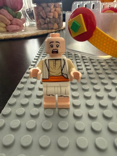 The scene depicts an indoor, domestic environment. The specific location within Limeil-Brévannes, France, is not discernible from the visual information provided.
A Lego minifigure stands centrally on a light grey Lego baseplate. The minifigure features a light nougat complexion, with a printed facial expression showing wide eyes and an open mouth. It is attired in a tan tunic with a dark brown collar, a white vest with vertical stripes, white trousers with a jagged black pattern at the lower edge, and an orange sash around the waist. Its light nougat hands are positioned slightly forward.
To the right of the minifigure, partially obscured by a translucent plastic container or barrier, is a yellow Lego arm element connected to a red circular Lego piece, which in turn features a yellow square Lego piece topped with a smaller green square Lego piece.
In the background, on the left, a dark brown surface, likely a table, is visible. On this surface, a clear glass jar contains numerous small, irregularly shaped confectionery items or nuts in shades of brown, grey, and orange. To the left of the jar, a cluster of pastel-colored candies (pink, green, yellow) rests on a light-colored woven mat. The background elements are partially out of focus.