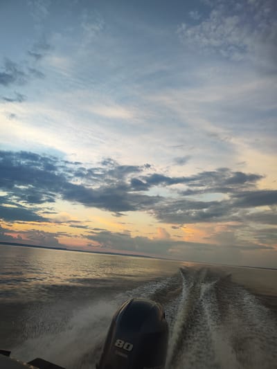 The image depicts a view from a boat moving through calm water at sunset. The sky is filled with a mix of light and dark clouds, with hues of orange, pink, and blue visible, suggesting the sun is setting or has recently set. The water is reflecting the colors of the sky and shows a prominent wake trailing behind the boat. In the foreground, the dark engine of the boat is visible, with the number "80" and the brand "YAMAHA" clearly discernible. The horizon line is flat, indicating a large body of water, possibly a lake or a wide river, with a distant landmass barely visible. The scene appears to be an outdoor activity, likely on a body of water. Without additional information, the specific location within the city of Галево, Russia cannot be determined from the visual elements of the image alone.